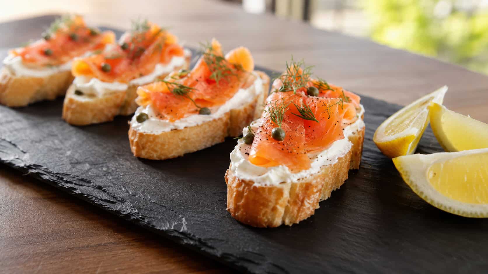 smoked salmon crostini topped with cream cheese, capers, and dill, lined up on a long slate board beside lemon wedges, in a wide horizontal shot, bright natural light, shallow depth of field, landscape orientation