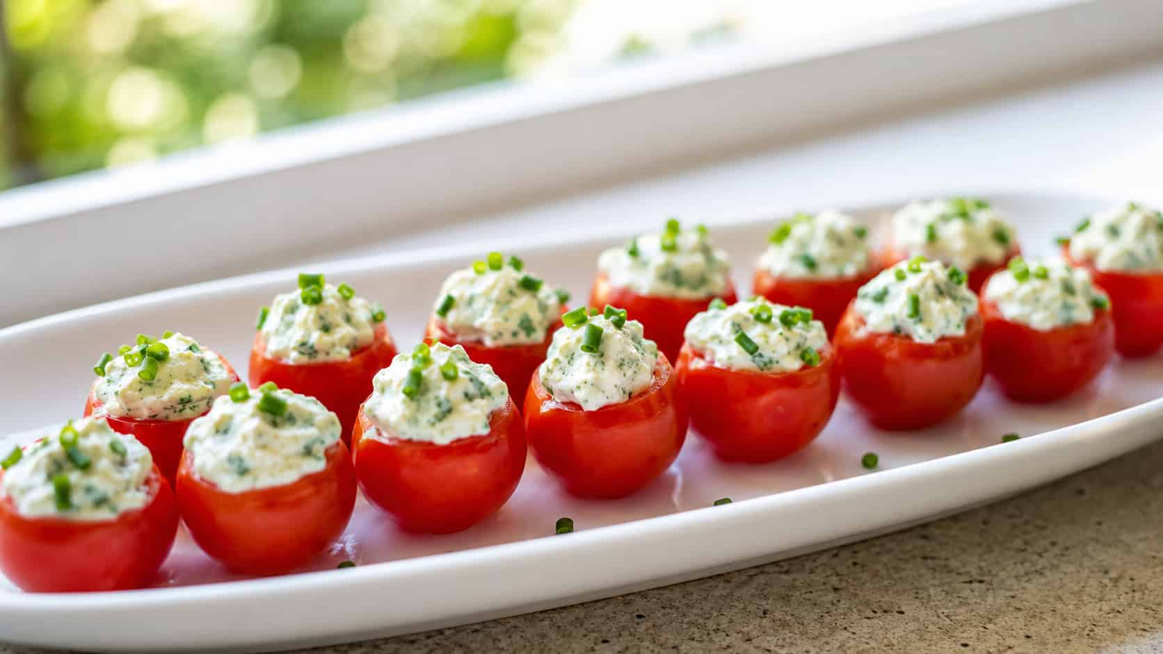cherry tomatoes stuffed with creamy herbed filling and sprinkled with chives, lined up on a white platter, in a wide horizontal shot, bright natural light, shallow depth of field, landscape orientation