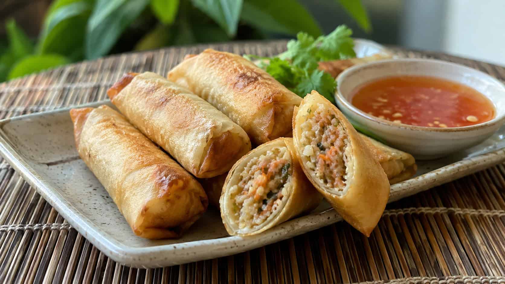 a wide horizontal platter of golden crispy pork spring rolls, one cut open to show the filling, with a bowl of sweet chili dipping sauce, on a bamboo mat, natural light, shallow depth of field, landscape orientation