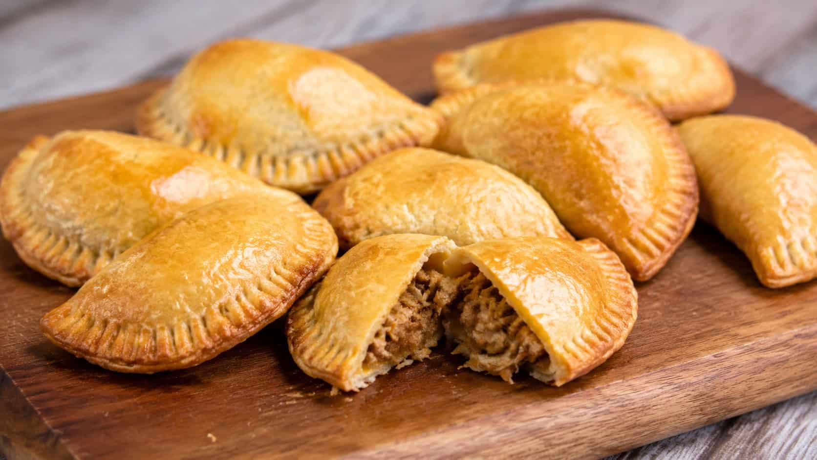 a wide horizontal arrangement of golden pork empanadas with crimped edges and shiny tops, one broken open to show the filling, on a wooden board, natural light, shallow depth of field, landscape orientation