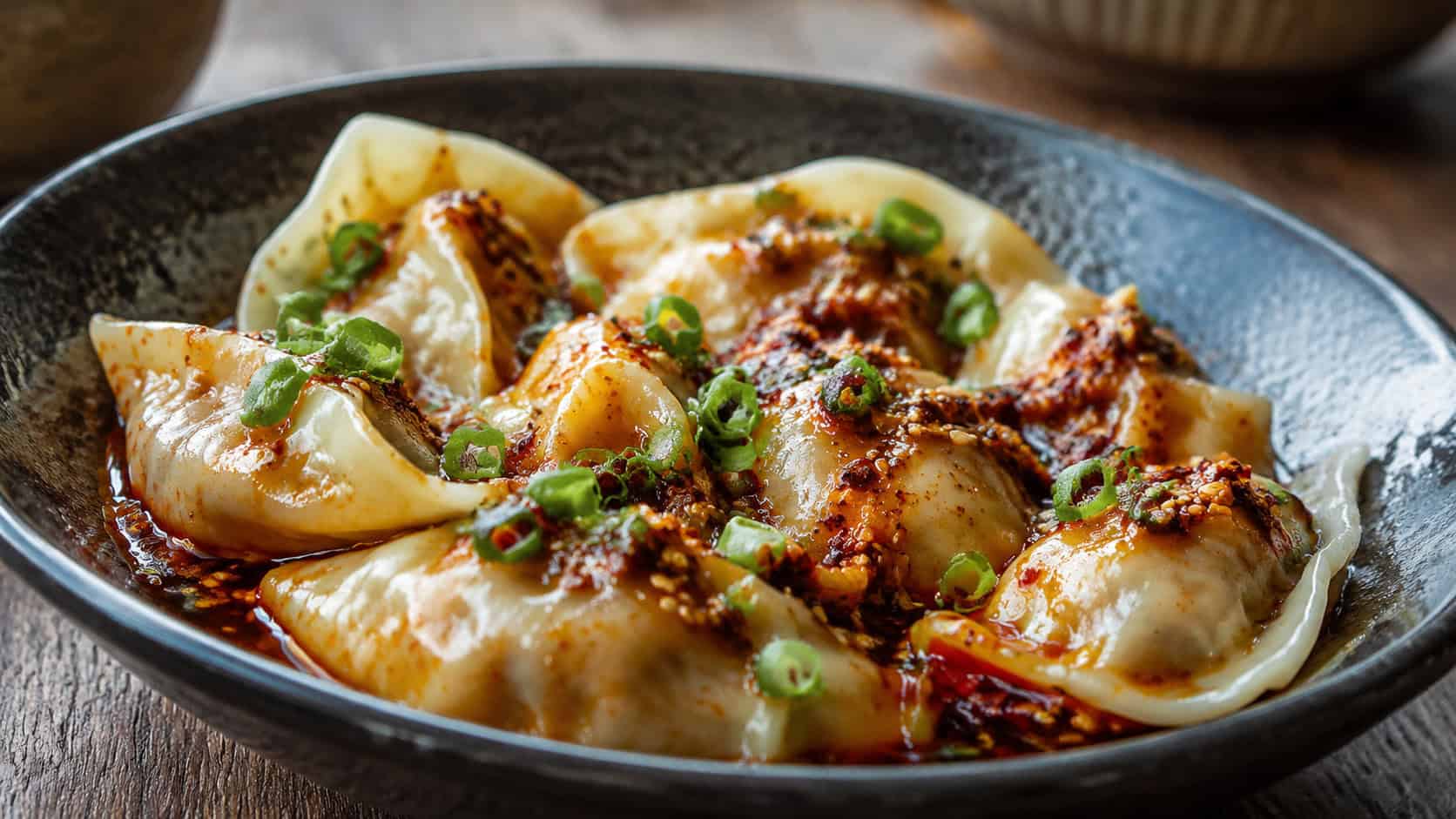 a wide horizontal bowl of plump Szechuan pork dumplings glistening in red chili oil, garnished with scallion and sesame seeds, on a dark ceramic dish, soft natural light, shallow depth of field, landscape orientation