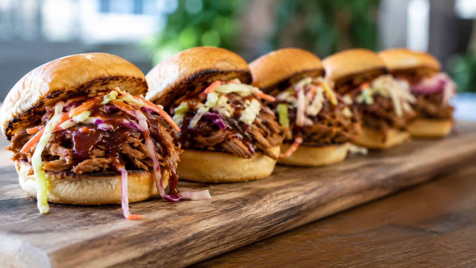 a wide horizontal row of mini pulled pork sliders on toasted buns with bright slaw spilling out, glossy barbecue sauce, lined up on a wooden serving board, natural light, shallow depth of field, landscape orientation