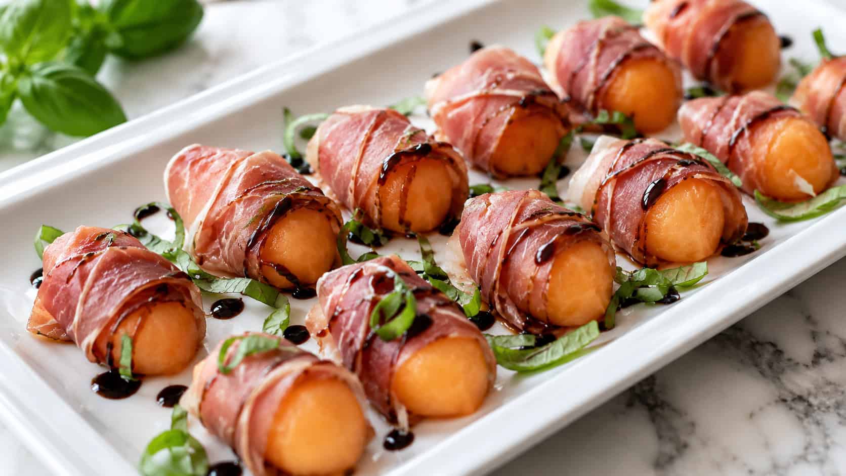 a wide horizontal platter of prosciutto-wrapped melon balls drizzled with dark balsamic, fresh basil scattered around, on a marble surface, bright natural light, shallow depth of field, landscape orientation