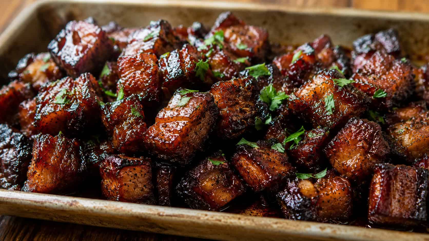 a wide horizontal pan of glistening pork belly burnt ends with deep caramelized edges and sticky glaze, sprinkled with chopped parsley, on a rustic metal tray, warm natural light, shallow depth of field, landscape orientation