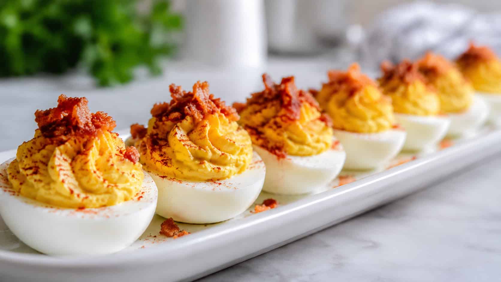 Creole deviled eggs lined up in a wide horizontal row on a white platter, creamy piped yellow filling dusted with paprika and topped with bacon bits, fresh bright light, shallow depth of field, landscape orientation