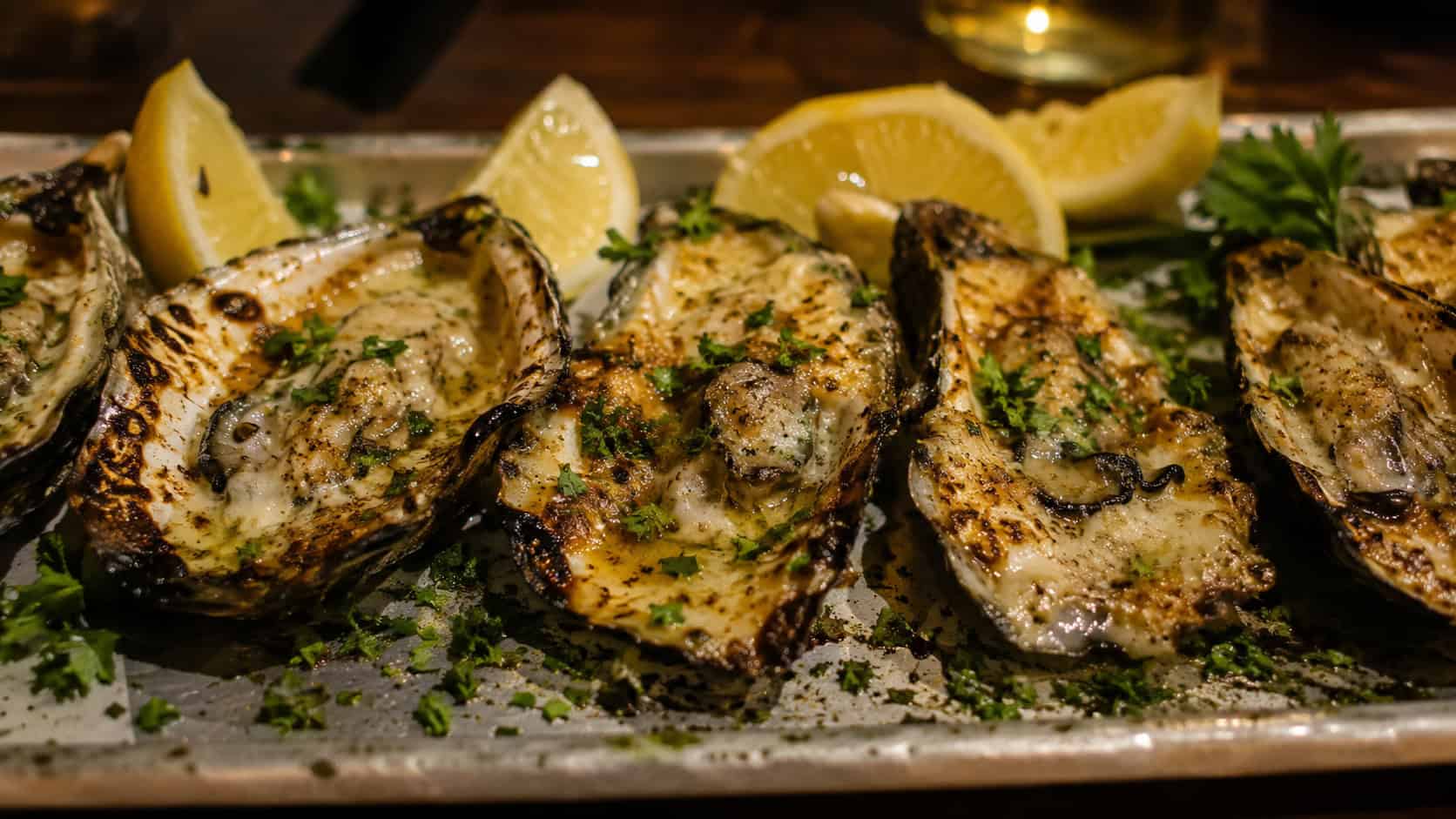 charbroiled oysters on the half shell arranged in a wide horizontal row on a metal tray, bubbling garlic butter and melted parmesan with charred edges, lemon wedges and parsley scattered around, moody warm light, shallow depth of field, landscape orientation