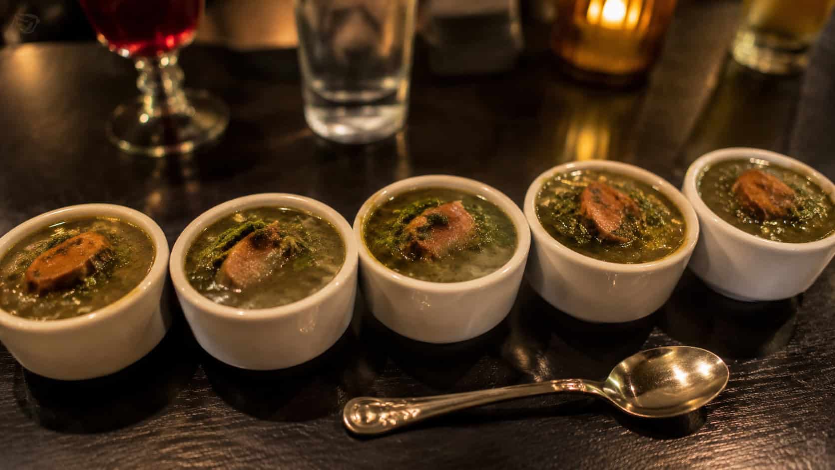 small cups of deep green gumbo z'herbes arranged in a wide horizontal line on a dark table, glossy greens and slices of smoked sausage in rich broth, a spoon resting nearby, warm low light, shallow depth of field, landscape orientation