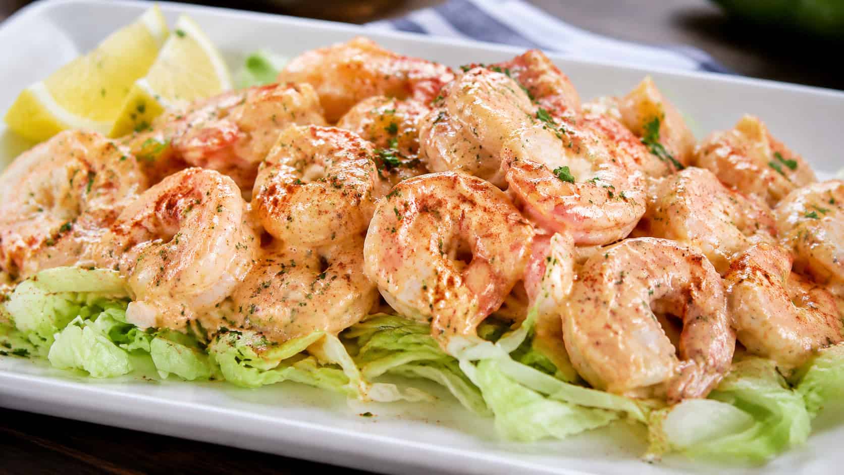 shrimp remoulade served over shredded lettuce in a wide horizontal frame on a white platter, glossy pink shrimp coated in creamy reddish sauce, lemon wedges and paprika dusting, bright fresh light, shallow depth of field, landscape orientation