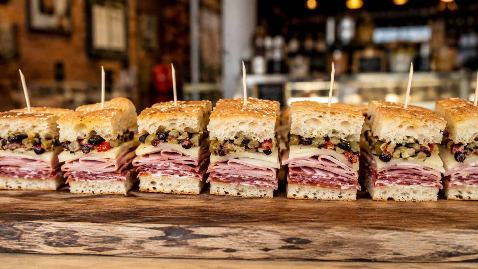 muffuletta bite squares arranged in a wide horizontal row on a wooden board, visible layers of cured meats cheese and olive salad, each speared with a toothpick, warm deli-style light, shallow depth of field, landscape orientation