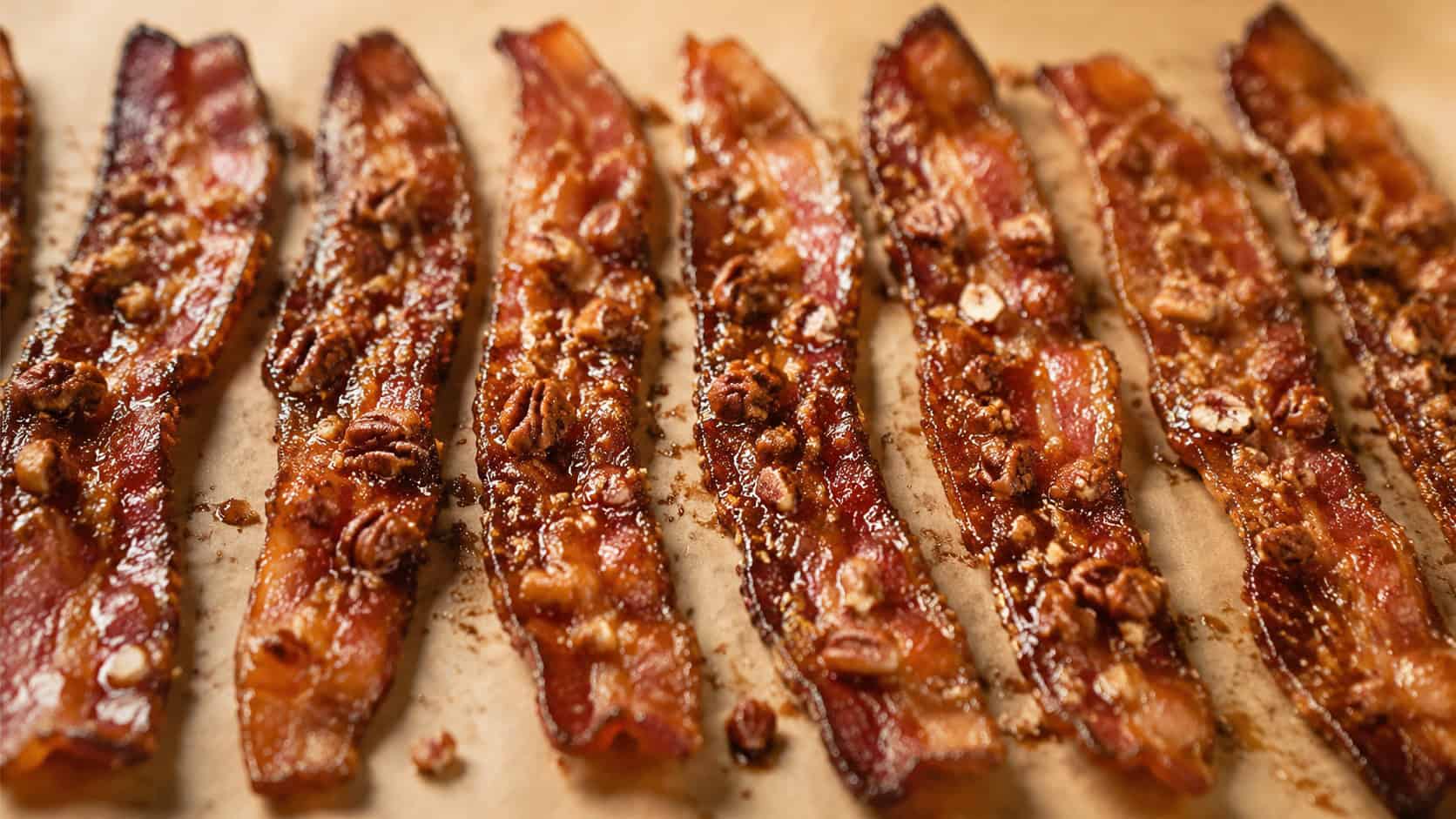candied pecan praline bacon strips arranged in a wide horizontal row on parchment, glossy caramelized brown sugar and chopped pecans clinging to the crispy bacon, warm cozy light, shallow depth of field, landscape orientation