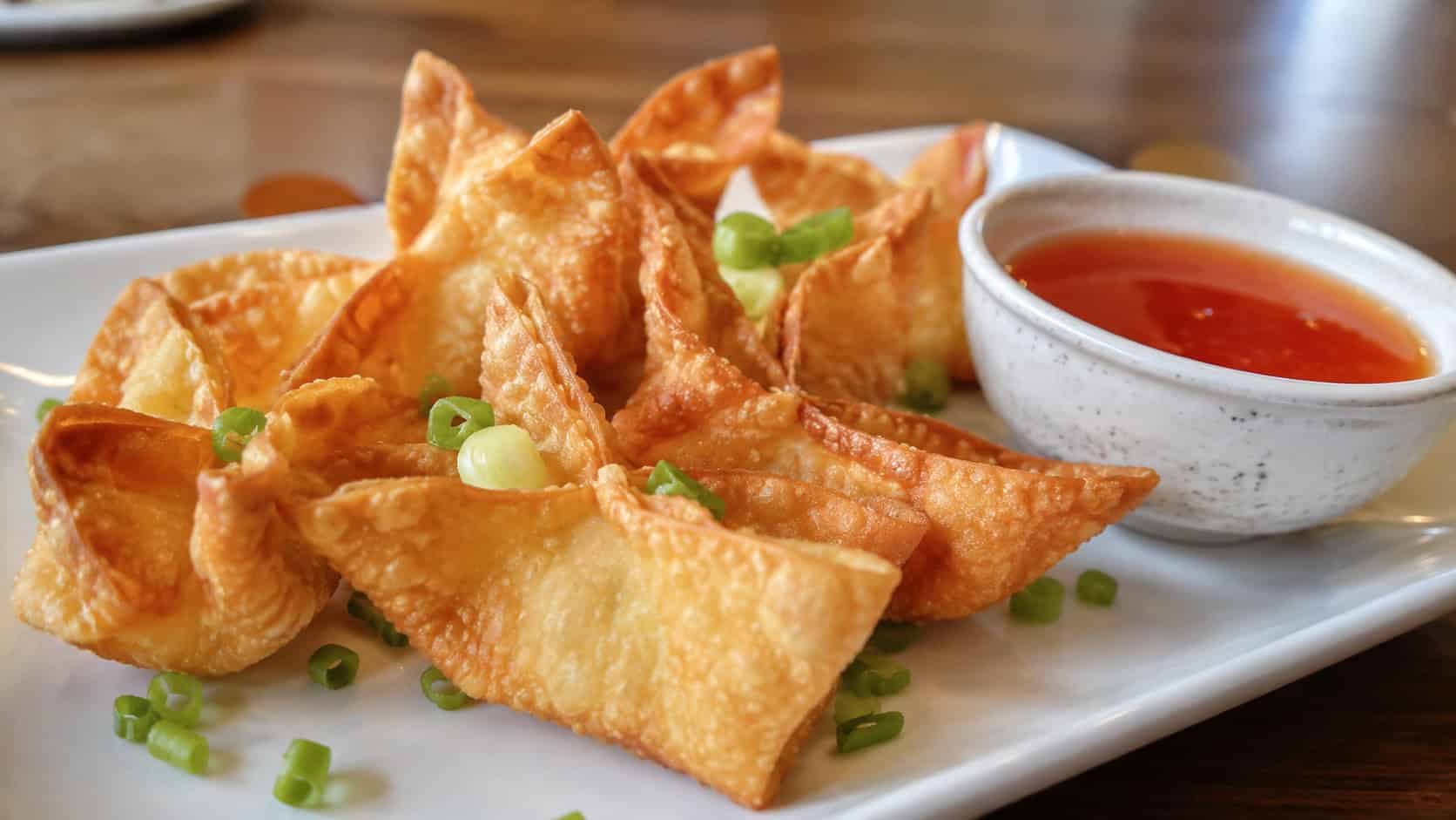 crispy golden crab rangoon in a wide horizontal shot, arranged on a plate with a bowl of sweet and sour sauce and green onion, natural light, shallow depth of field, landscape orientation