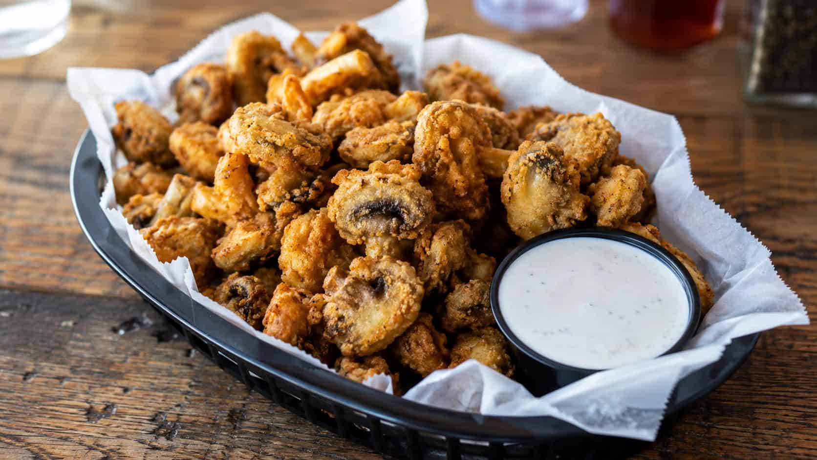 crispy fried mushrooms in a wide horizontal shot, golden and piled in a basket with a bowl of ranch on a rustic table, natural light, shallow depth of field, landscape orientation
