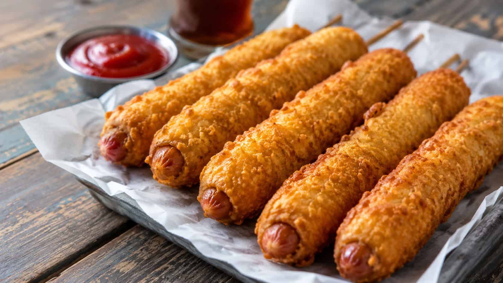crispy battered sausages in a wide horizontal shot, golden and lined up on a paper-lined tray with ketchup on a rustic table, natural light, shallow depth of field, landscape orientation