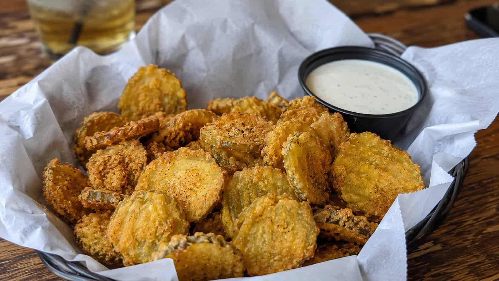 crispy fried pickle chips in a wide horizontal shot, piled in a basket lined with paper beside a bowl of ranch on a rustic table, natural light, shallow depth of field, landscape orientation