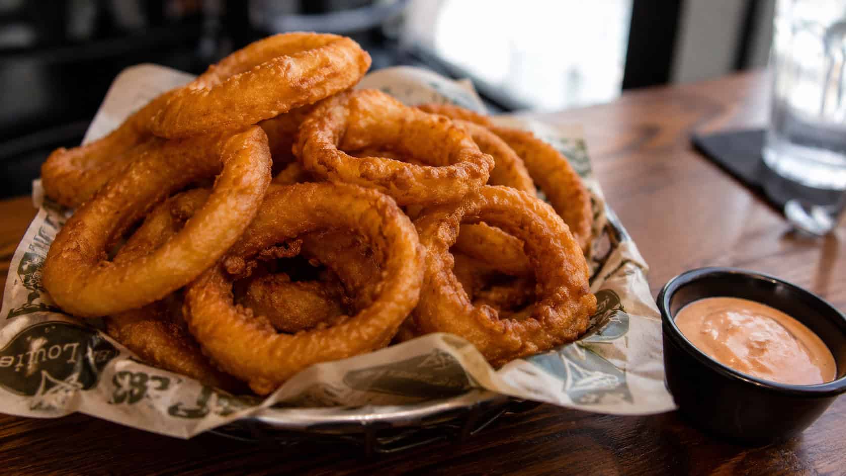 golden crispy onion rings in a wide horizontal shot, stacked high in a basket with a bowl of dipping sauce on a wooden table, natural light, shallow depth of field, landscape orientation