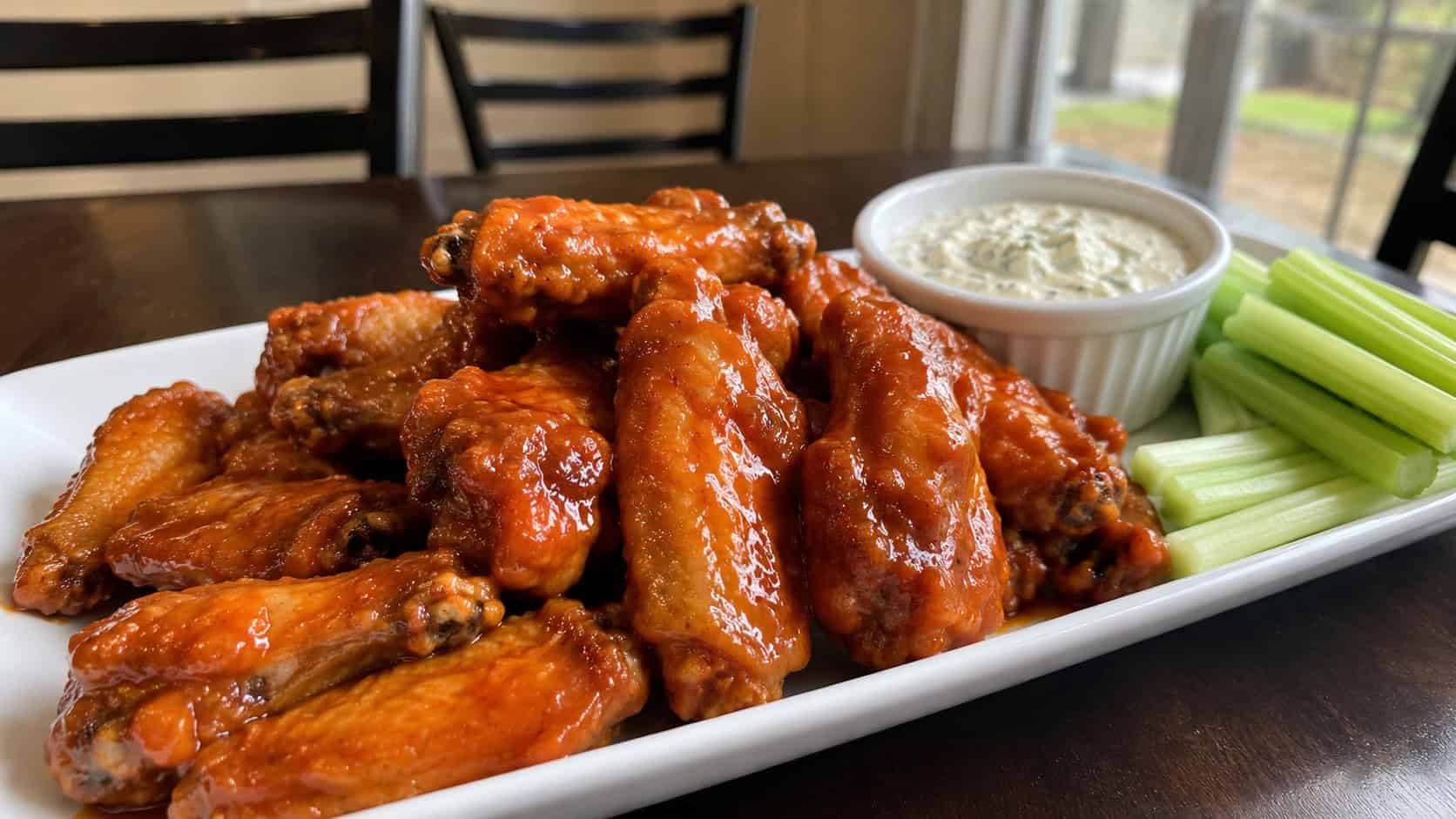 glossy buffalo chicken wings in a wide horizontal shot, piled on a platter with celery sticks and blue cheese dip, natural light, shallow depth of field, landscape orientation
