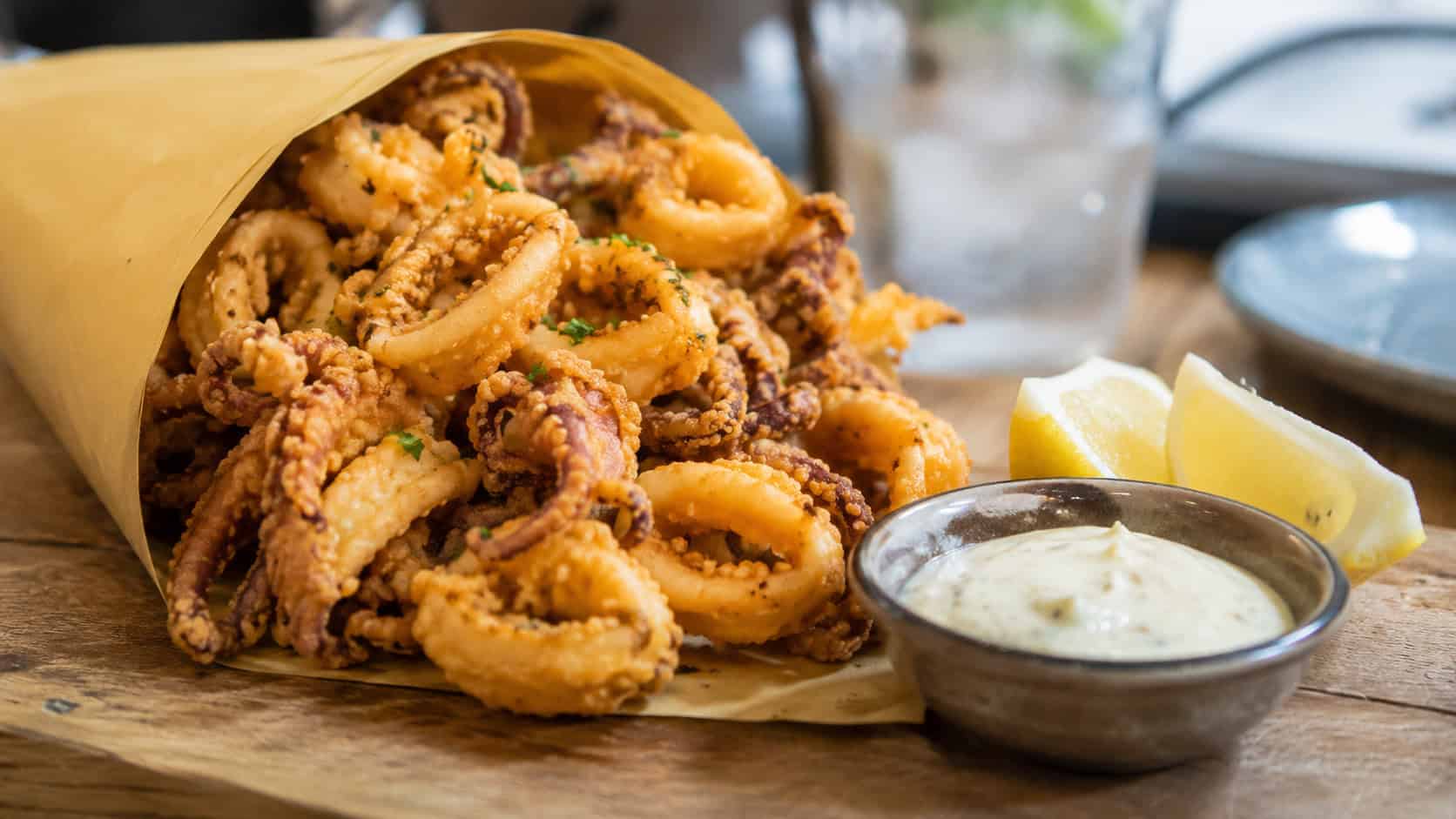 crispy fried calamari in a wide horizontal shot, piled in a paper cone with lemon wedges and dipping sauce on a wooden table, natural light, shallow depth of field, landscape orientation