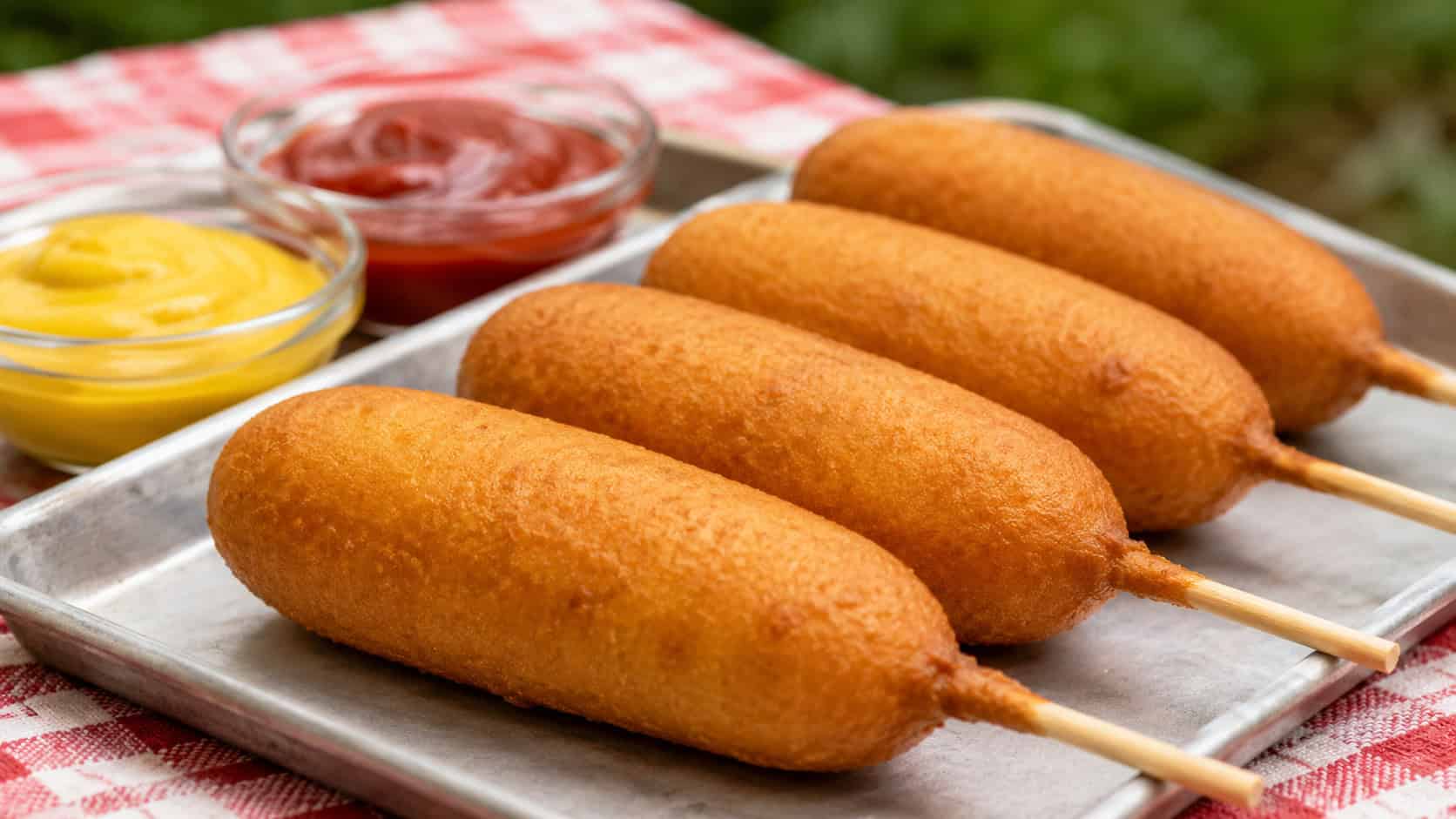 golden corn dogs on sticks in a wide horizontal shot, lined up on a tray with mustard and ketchup on a checkered cloth, natural light, shallow depth of field, landscape orientation