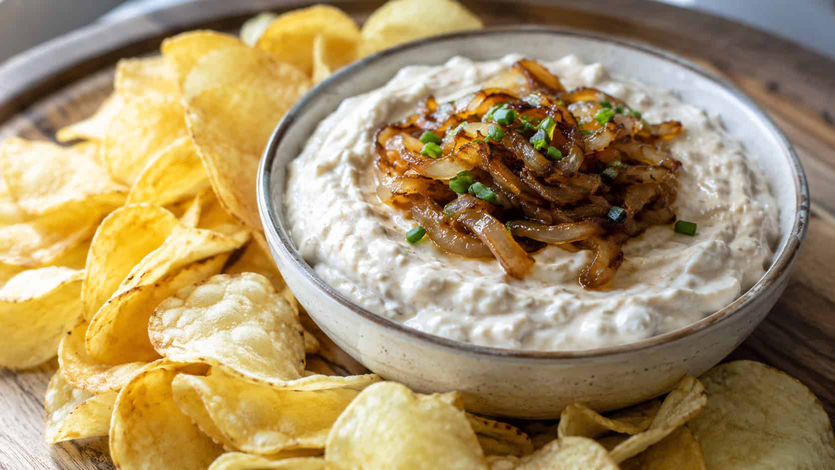 creamy Maui onion dip in a bowl topped with caramelized onion, in a wide horizontal shot, surrounded by kettle chips, on a wooden serving board, natural light, shallow depth of field, landscape orientation
