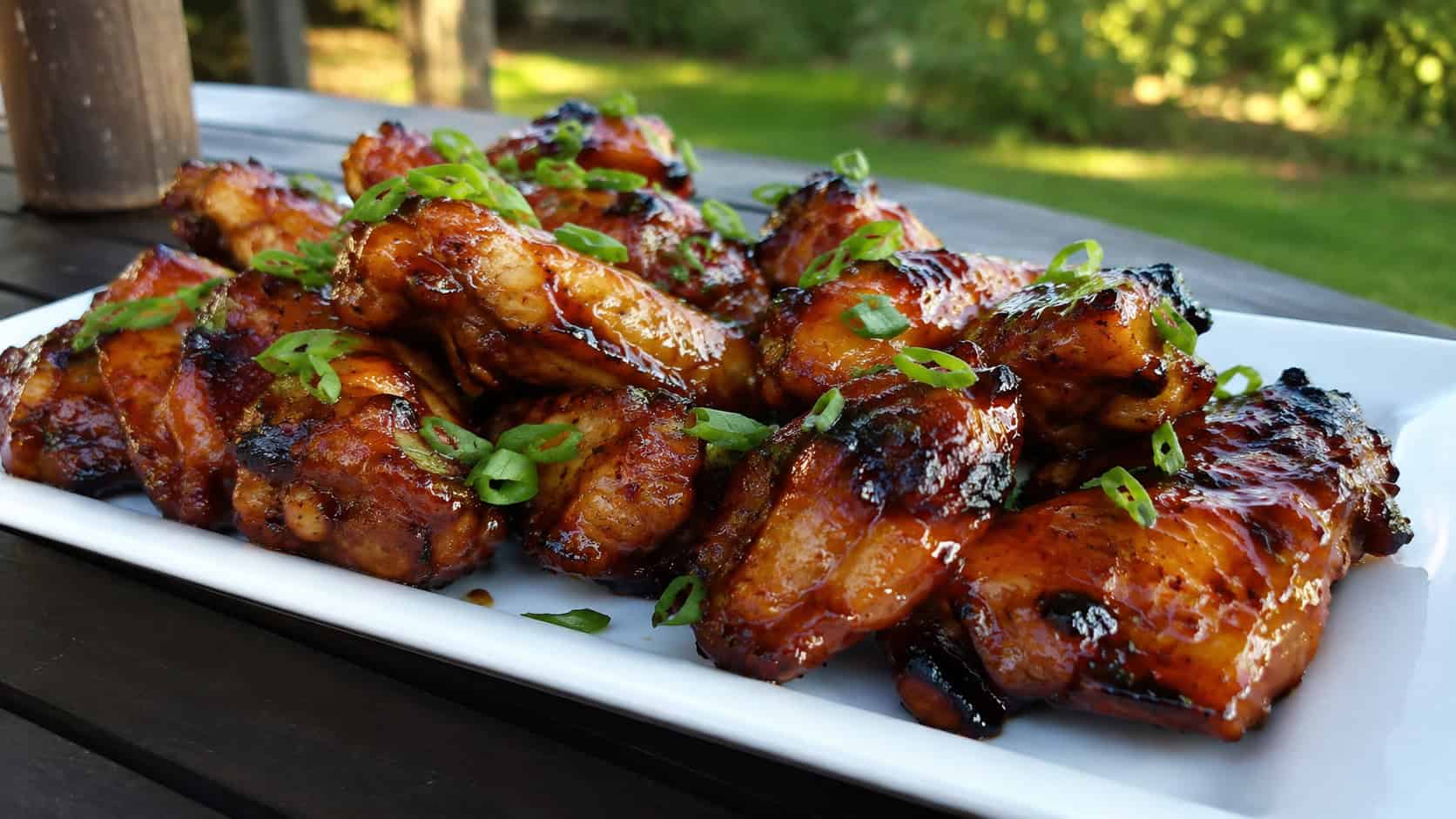 sticky huli huli chicken wings piled on a platter, in a wide horizontal shot, glossy caramelized glaze, garnished with green onion, on an outdoor table, warm natural light, shallow depth of field, landscape orientation