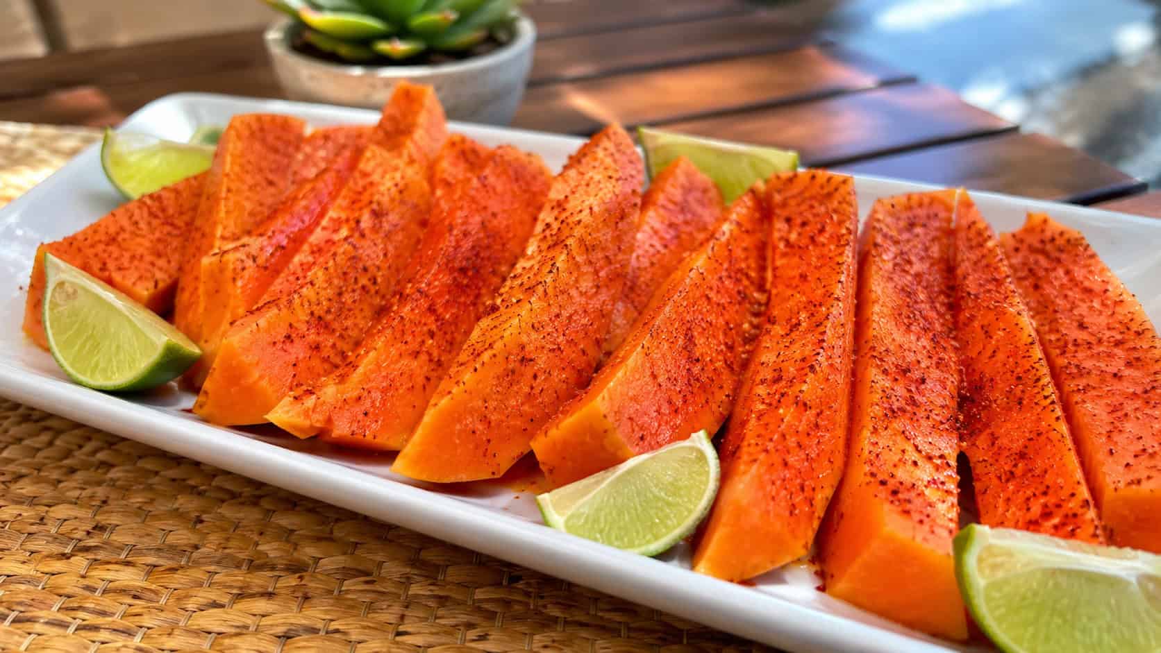 fresh papaya spears dusted with red Tajin and lime, in a wide horizontal shot, vibrant orange fruit on a white platter with lime wedges, on a sunny table, vivid natural light, shallow depth of field, landscape orientation
