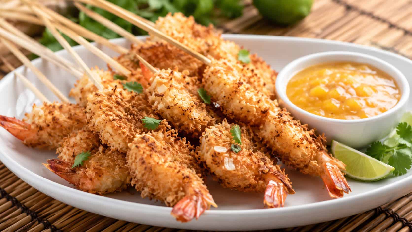 crispy coconut shrimp skewers fanned out on a platter, in a wide horizontal shot, golden coconut coating with a bowl of mango dipping sauce, on a bamboo mat, bright natural light, shallow depth of field, landscape orientation