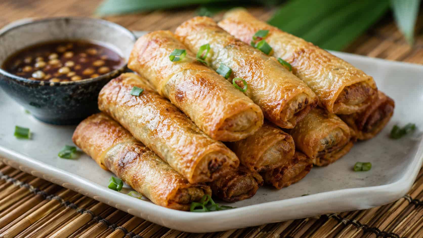 crispy kalua pork spring rolls stacked on a platter, in a wide horizontal shot, golden crunchy wrappers with a bowl of dipping sauce, on a bamboo mat, natural light, shallow depth of field, landscape orientation