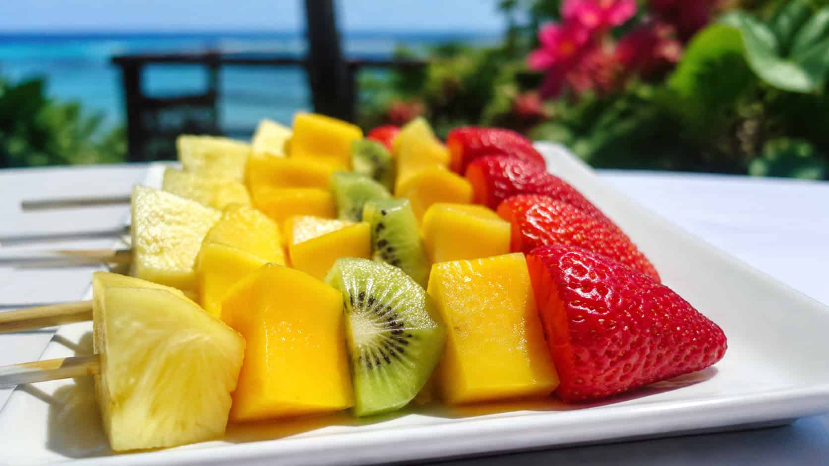 colorful tropical fruit skewers arranged on a white platter, in a wide horizontal shot, pineapple, mango, kiwi, and strawberries glistening, on a bright outdoor table, vivid natural light, shallow depth of field, landscape orientation