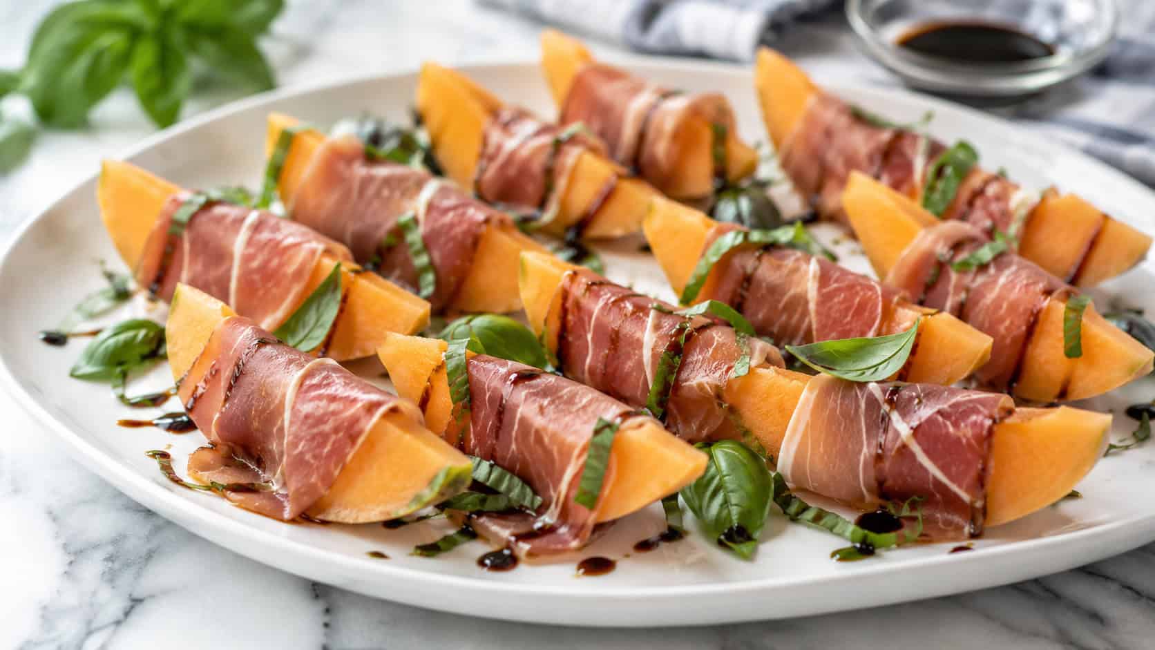 a wide platter of orange cantaloupe wedges wrapped in pink prosciutto with fresh basil, drizzled with balsamic glaze, on a marble surface, bright natural light, shallow depth of field, landscape orientation