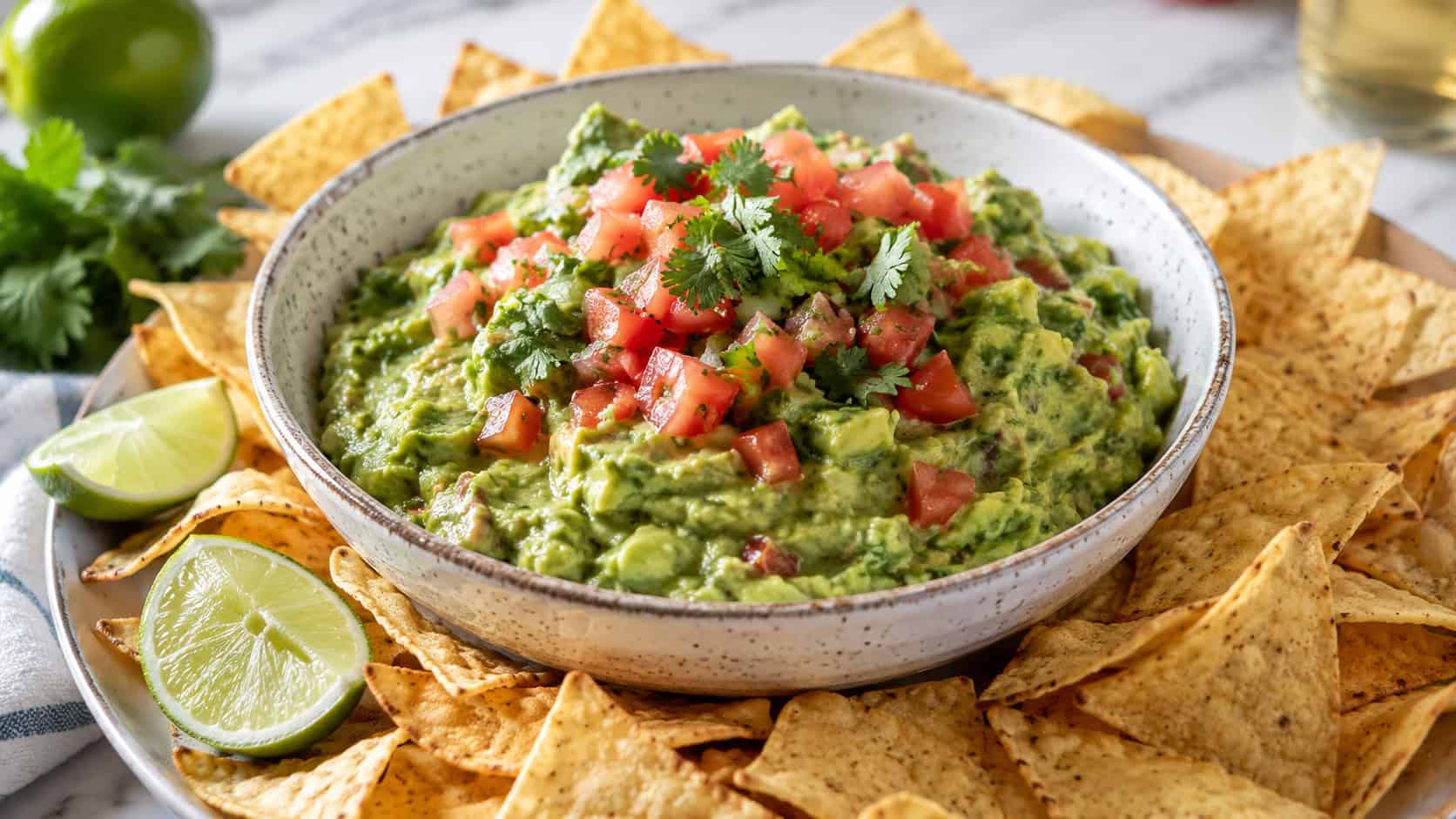 a wide bowl of chunky green guacamole topped with diced tomato and cilantro, surrounded by golden tortilla chips, lime wedges nearby, on a bright surface, natural light, shallow depth of field, landscape orientation