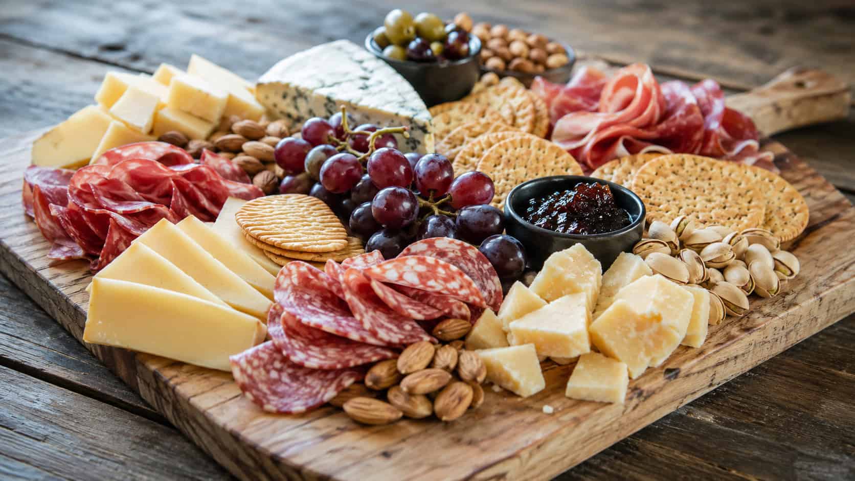 a wide wooden board loaded with assorted cheeses, folded cured meats, crackers, grapes, and nuts arranged in sections, on a rustic table, natural light, shallow depth of field, landscape orientation