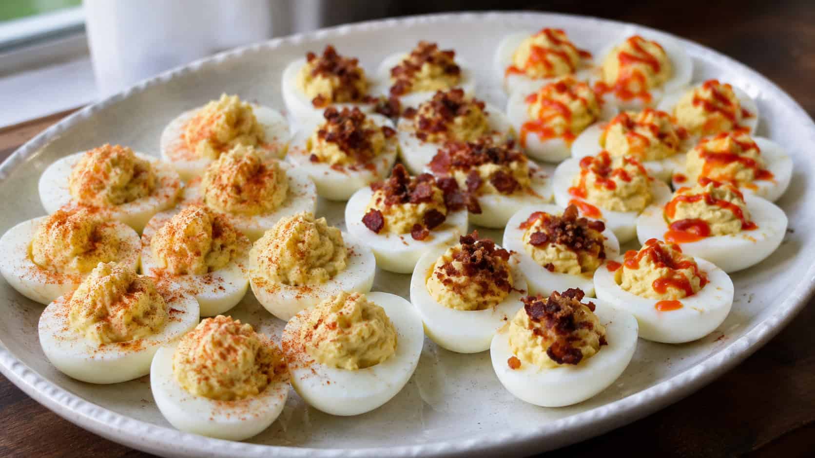 a wide platter of deviled eggs arranged in three sections with different garnishes of paprika, bacon, and red sriracha, on a light ceramic dish, natural light, shallow depth of field, landscape orientation