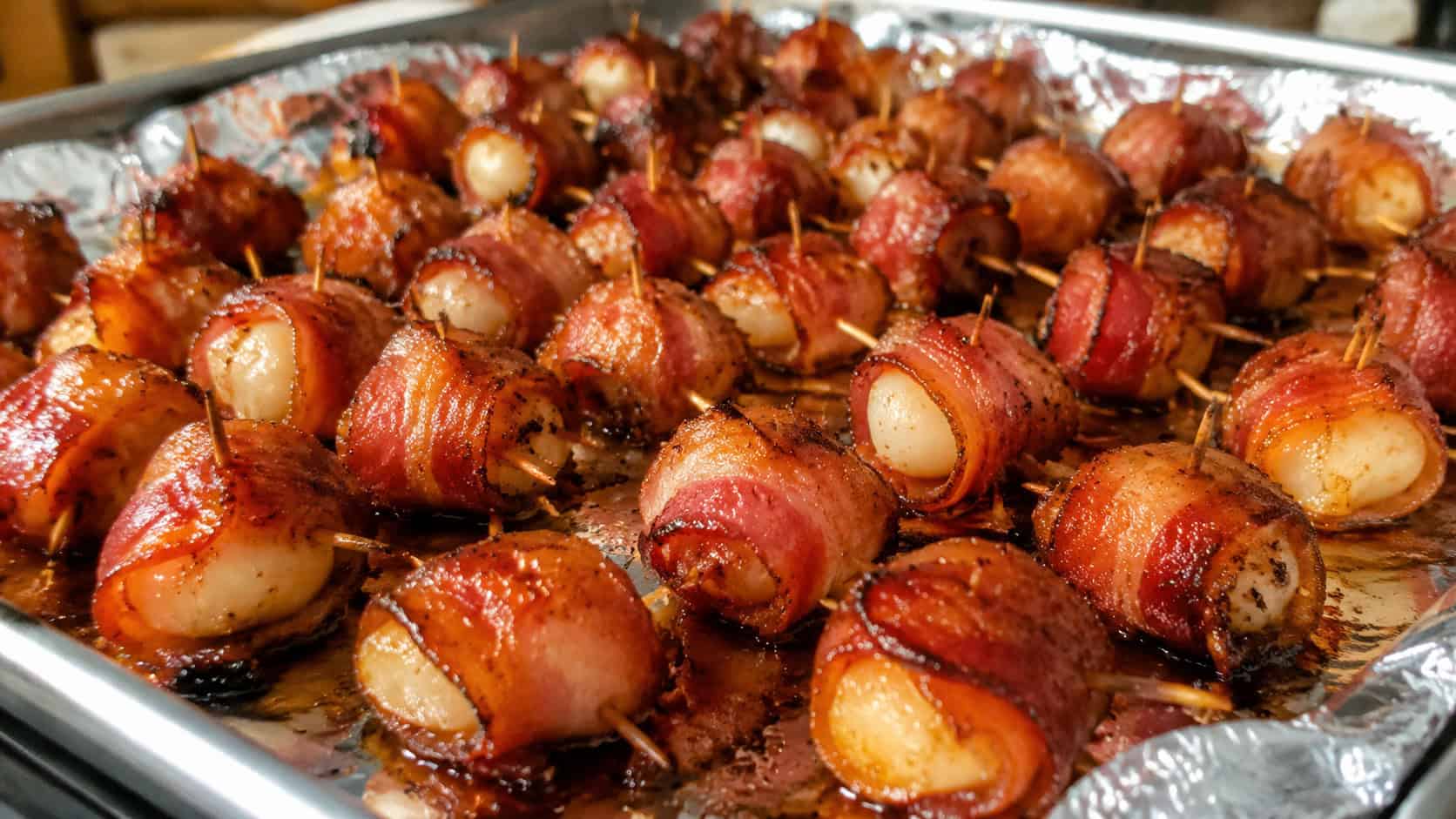 a wide tray of crispy bacon-wrapped water chestnuts glazed and glistening, secured with toothpicks, on a foil-lined sheet, warm natural light, shallow depth of field, landscape orientation