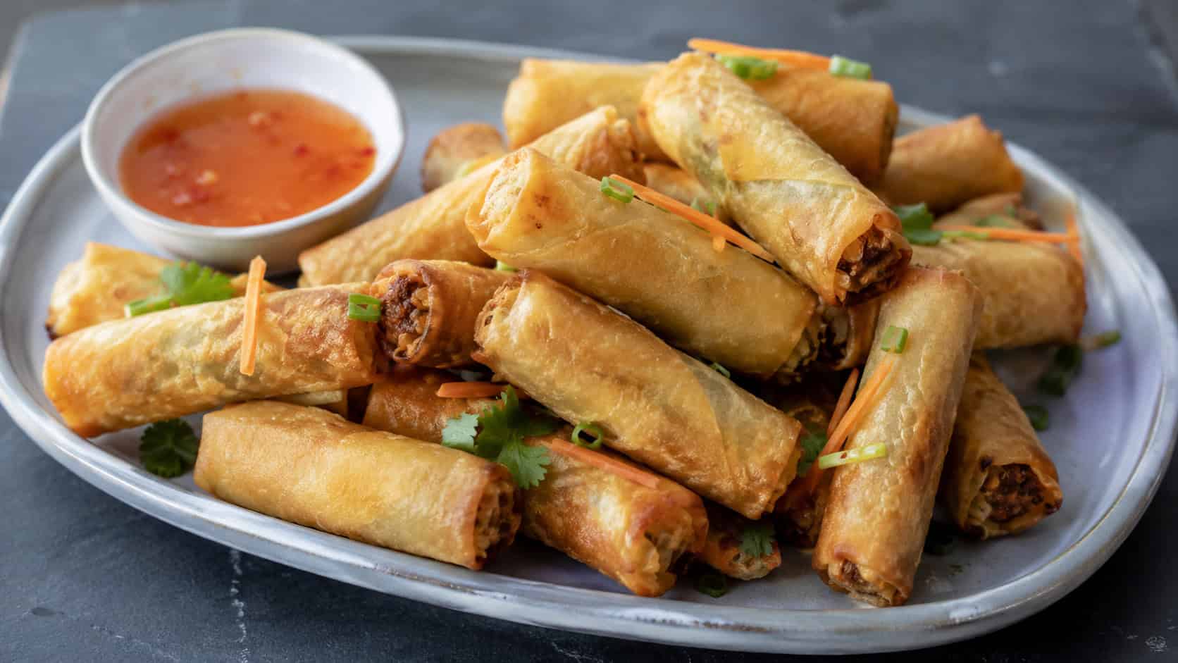 a wide platter of golden crispy vegetable spring rolls stacked loosely with a small bowl of dipping sauce, on a dark slate surface, natural light, shallow depth of field, landscape orientation