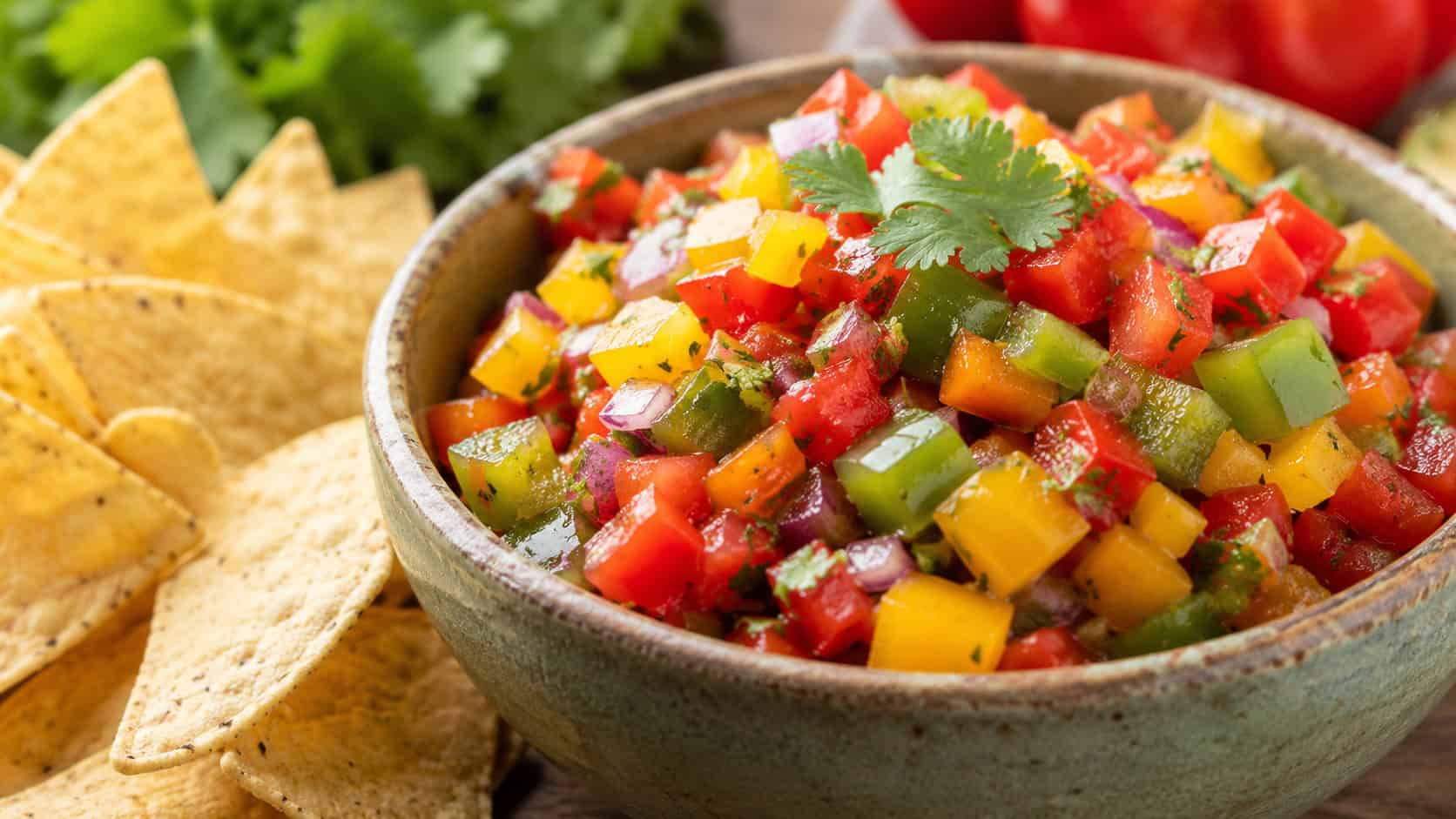 fresh chunky bell pepper salsa with red, yellow, and green peppers and cilantro in a rustic bowl beside tortilla chips, in a wide horizontal shot, bright natural light, shallow depth of field, landscape orientation