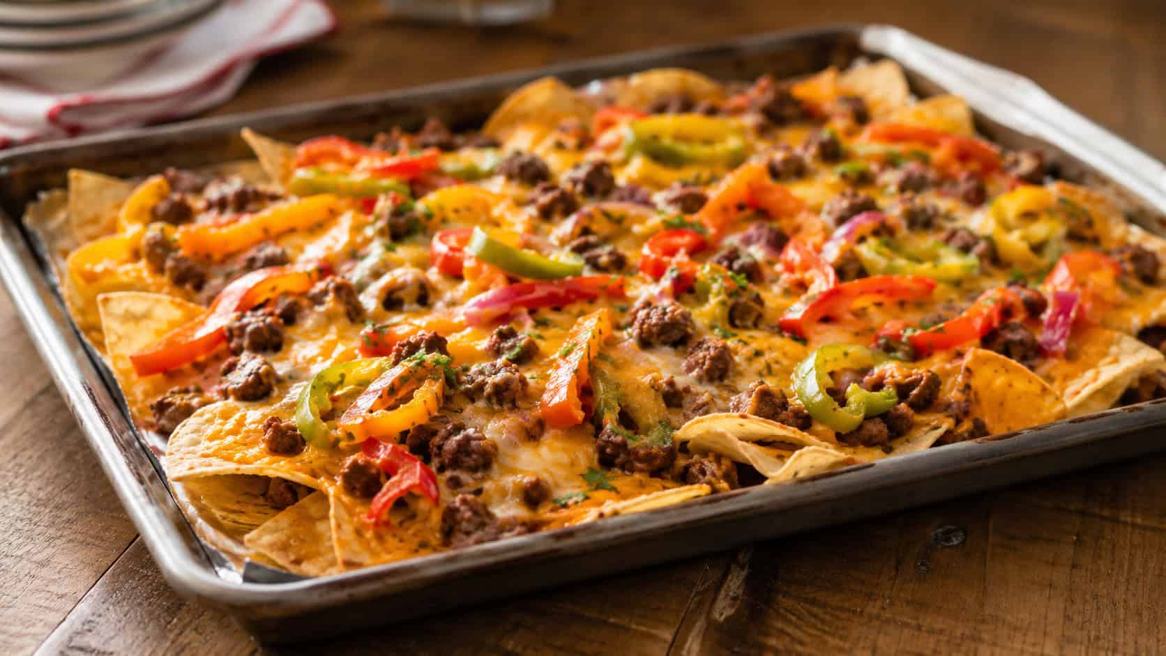 loaded sheet pan nachos with melted cheese, ground beef, and colorful sliced peppers, on a wooden table, in a wide horizontal shot, warm natural light, shallow depth of field, landscape orientation