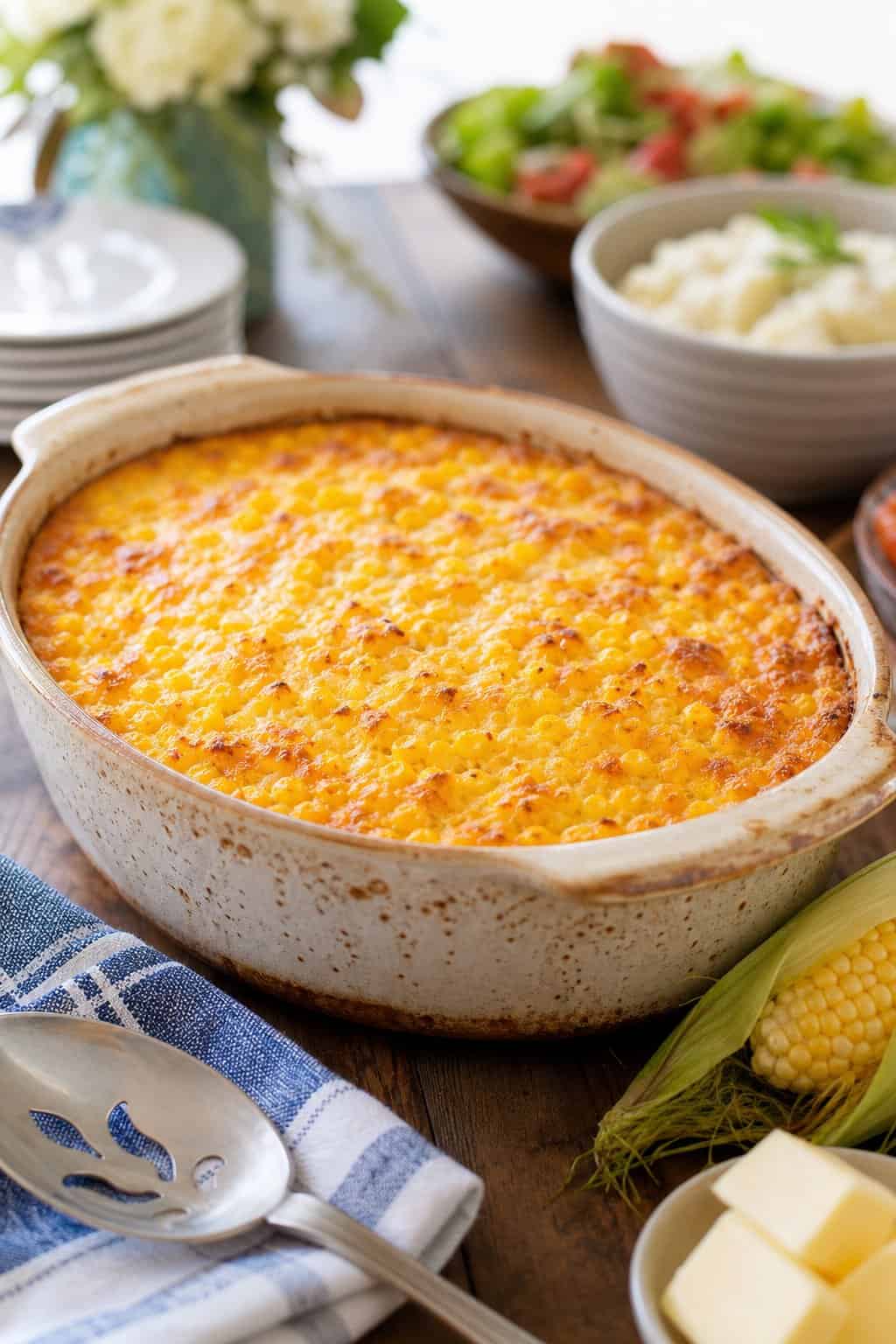 A golden-topped corn casserole baked in a white ceramic dish on a wooden table, with a large serving spoon and two ears of fresh corn arranged to the side.