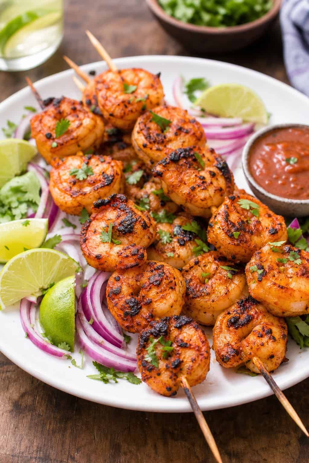 A plate of grilled chipotle-style shrimp skewers garnished with lime wedges, cilantro, and red onions on a wooden table.