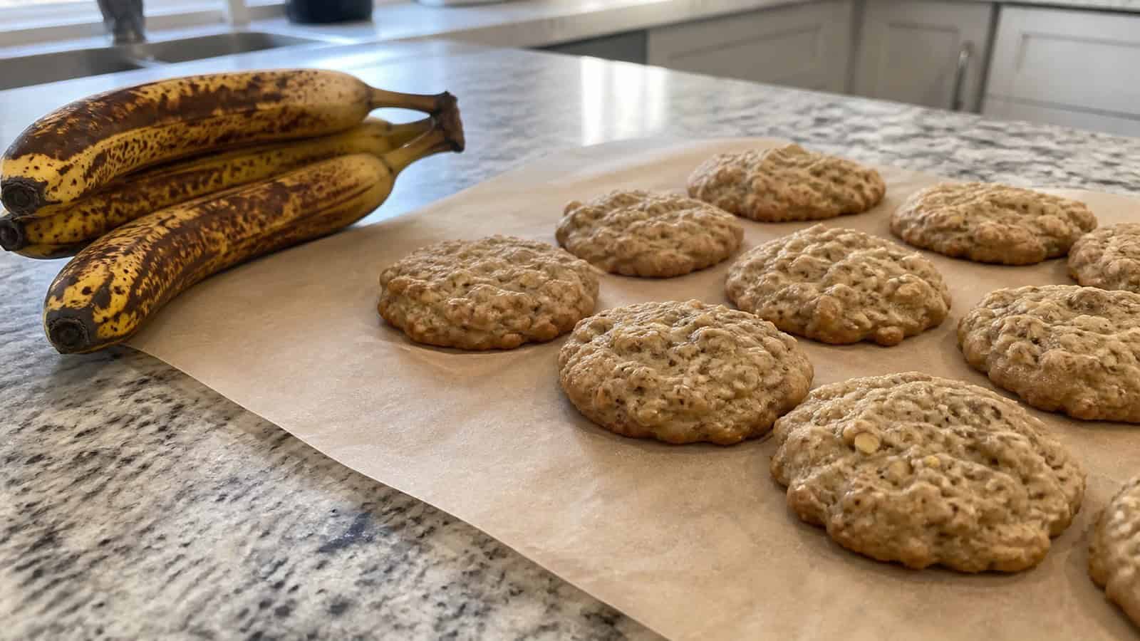 banana cookies cooling on parchment paper beside overripe bananas on a kitchen counter