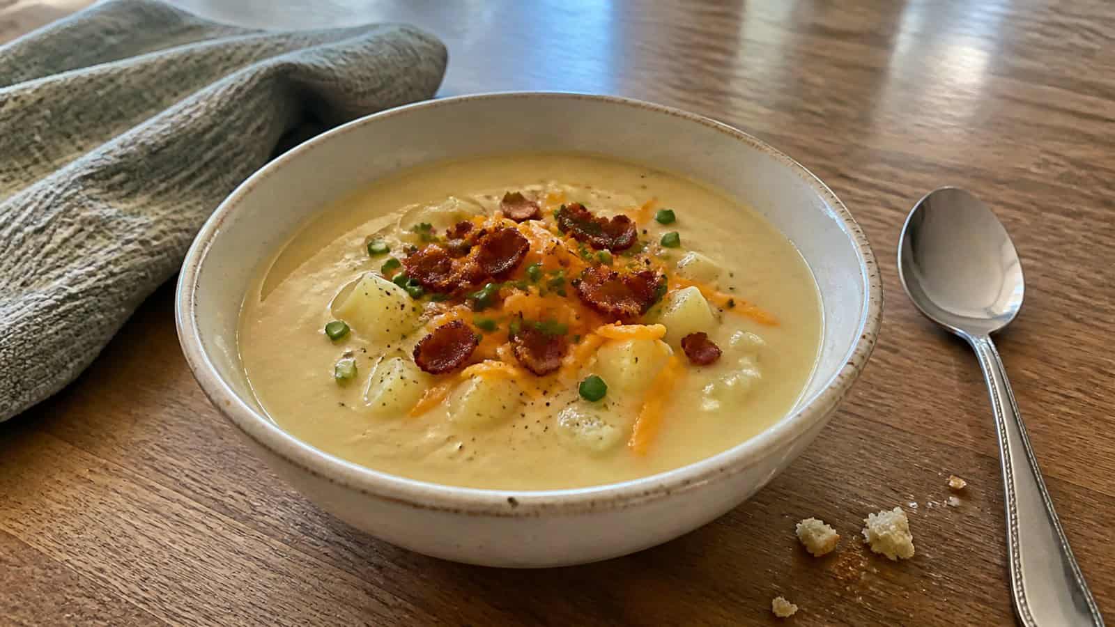 a bowl of homemade potato soup on a kitchen table with a spoon and a few crumbs nearby