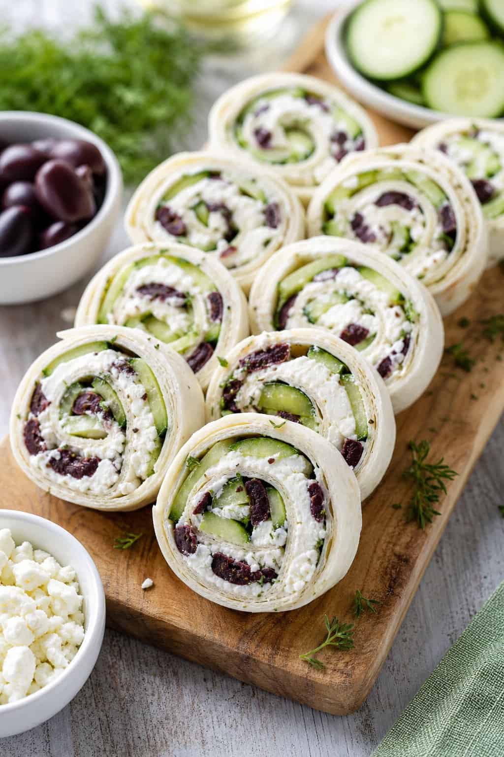 15 Pinwheel Appetizers for Game Day 18 Plate of Greek feta, cucumber, and olive pinwheel appetizers on a wooden serving board showing white crumbled feta, thin cucumber rounds, and dark kalamata olives inside sliced flour tortilla rolls, with small bowls of olives and feta nearby.