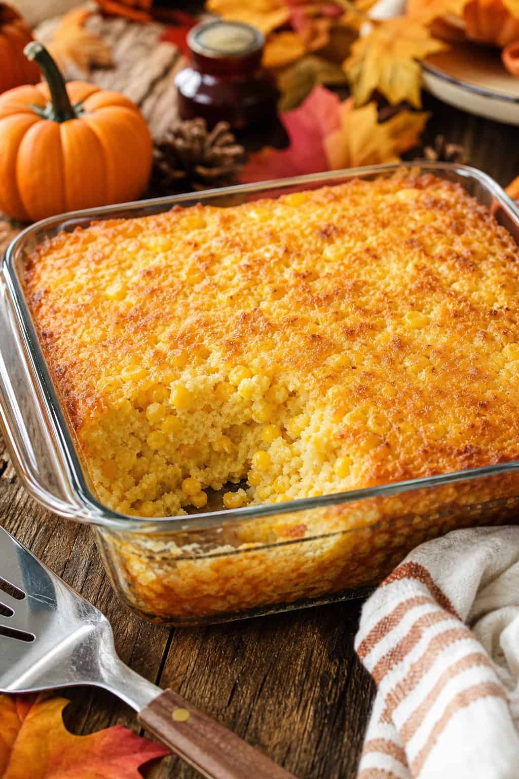 A golden-brown corn casserole in a clear glass baking dish on a wooden table with small decorative gourds and dried leaves arranged around it.