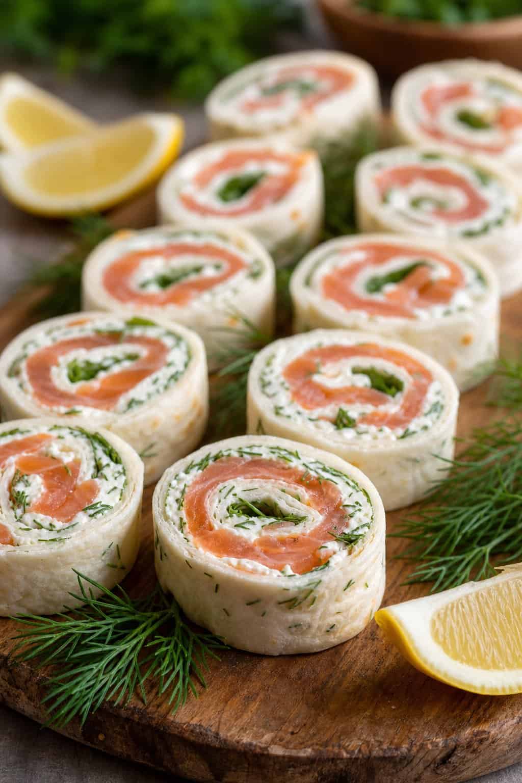 Close-up of smoked salmon dill cream cheese pinwheels on a wooden board showing pale pink salmon slices and thin cucumber rounds inside rolled tortillas, with fresh dill sprigs and lemon wedges alongside.