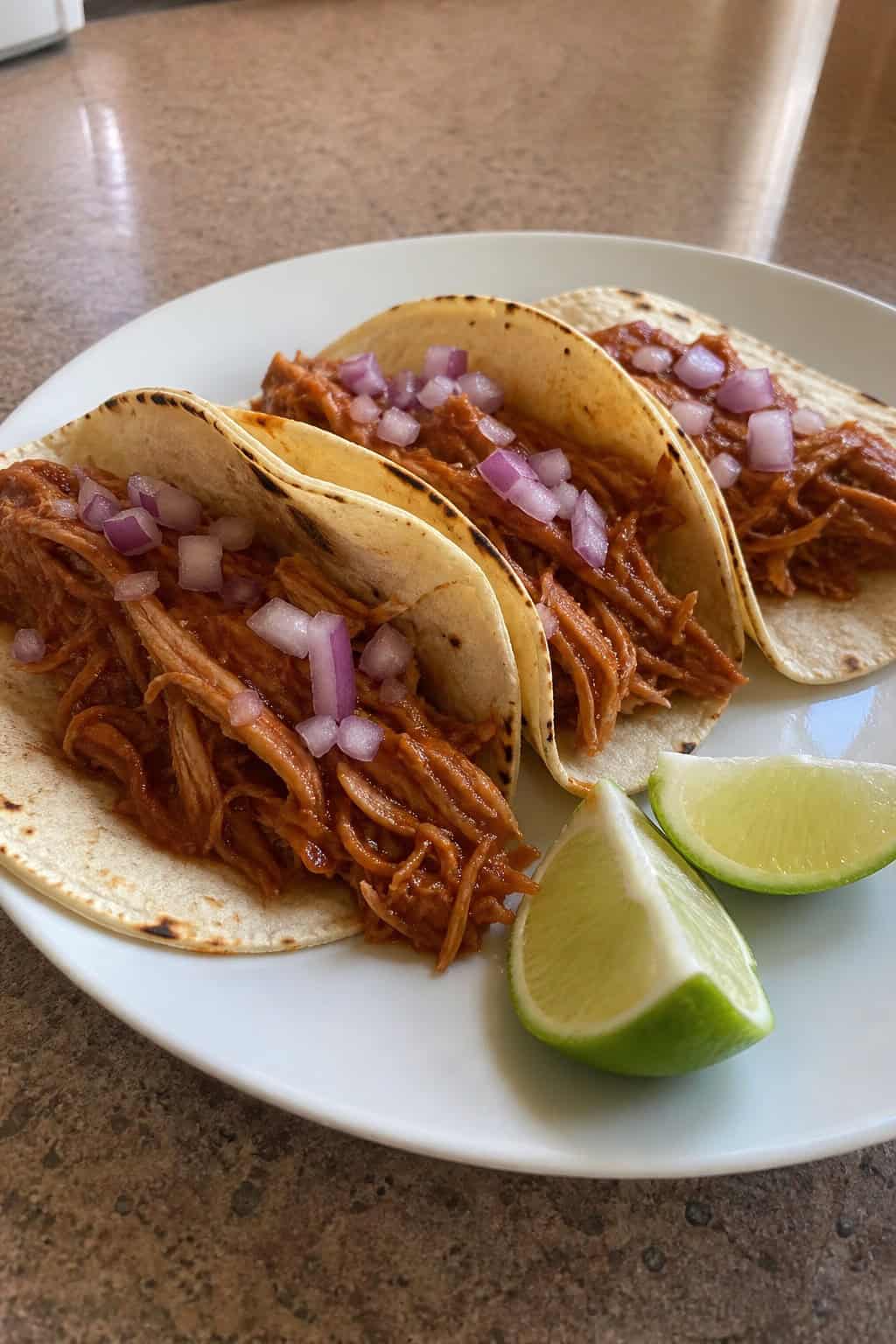 A wooden table with a plate of saucy BBQ shredded chicken tacos topped with fresh cilantro and diced red onion, lime wedges resting alongside.