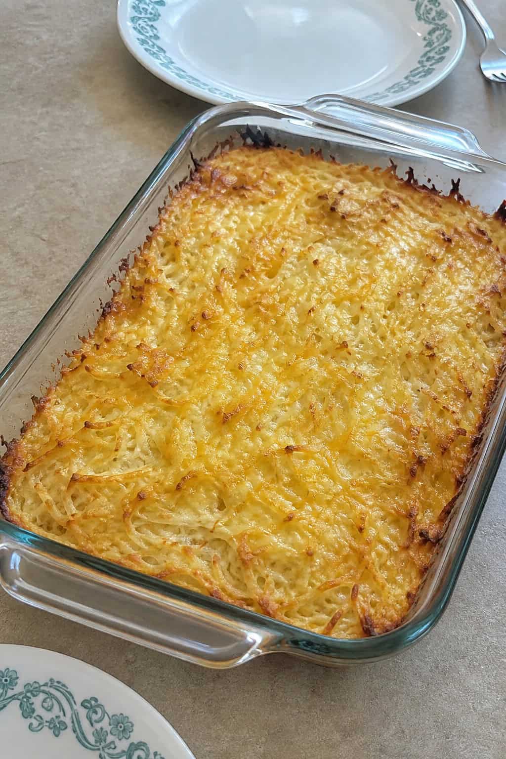 A freshly baked cheesy hashbrown casserole in a glass baking dish on a wooden table with a serving spatula and fresh ingredients nearby.