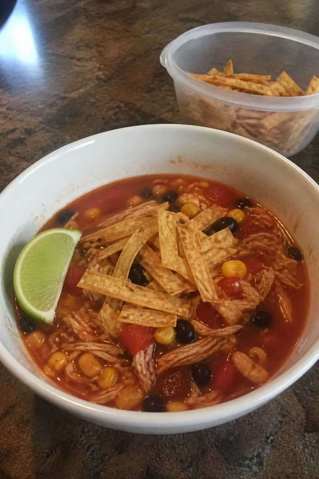 A bowl of chicken tortilla soup with shredded chicken, tomatoes, corn, beans, and tortilla strips on a wooden table.