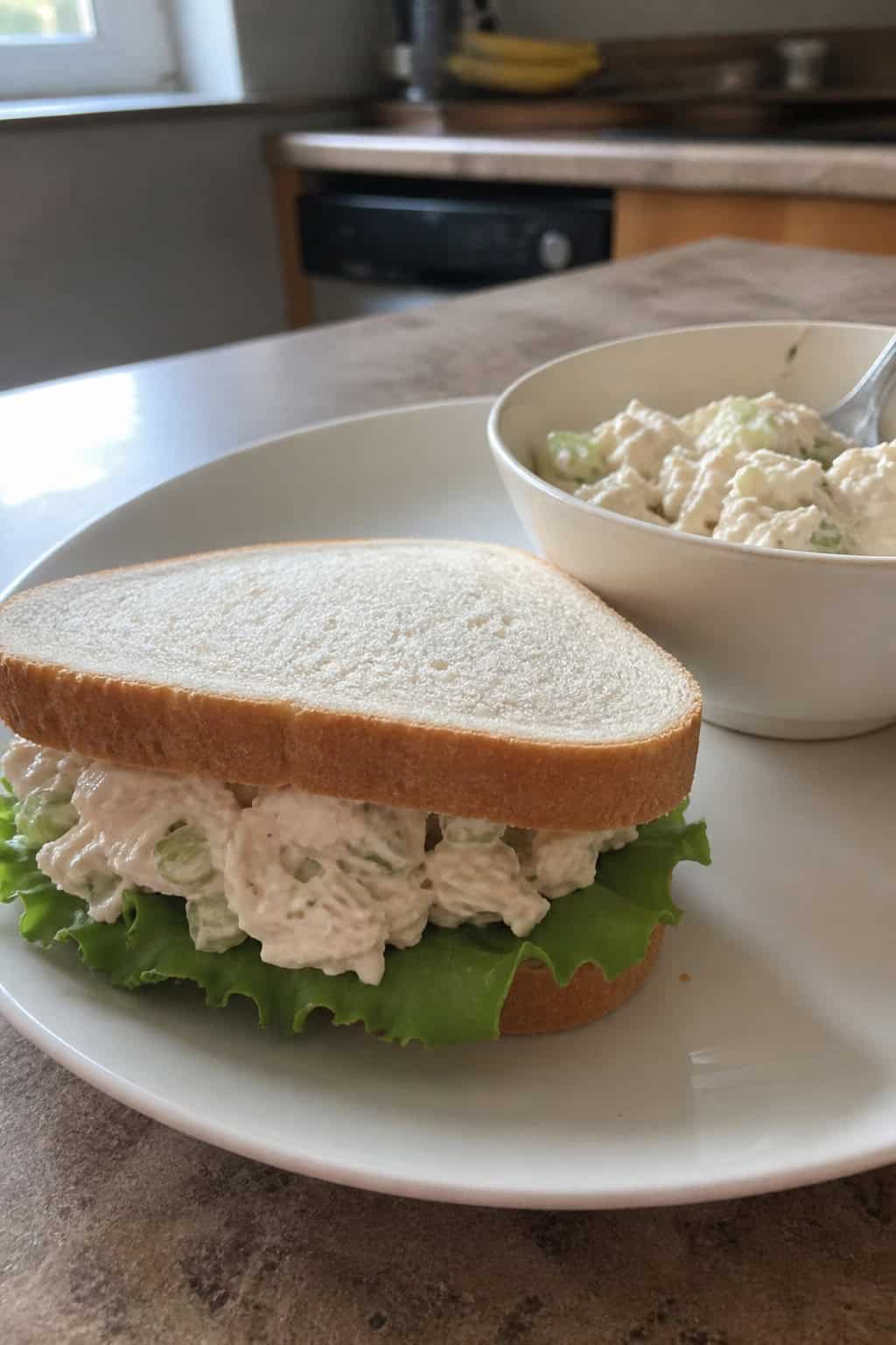 A chicken salad sandwich on a wooden board with fresh lettuce, tomato slices, and a bowl of chicken salad on a kitchen countertop.