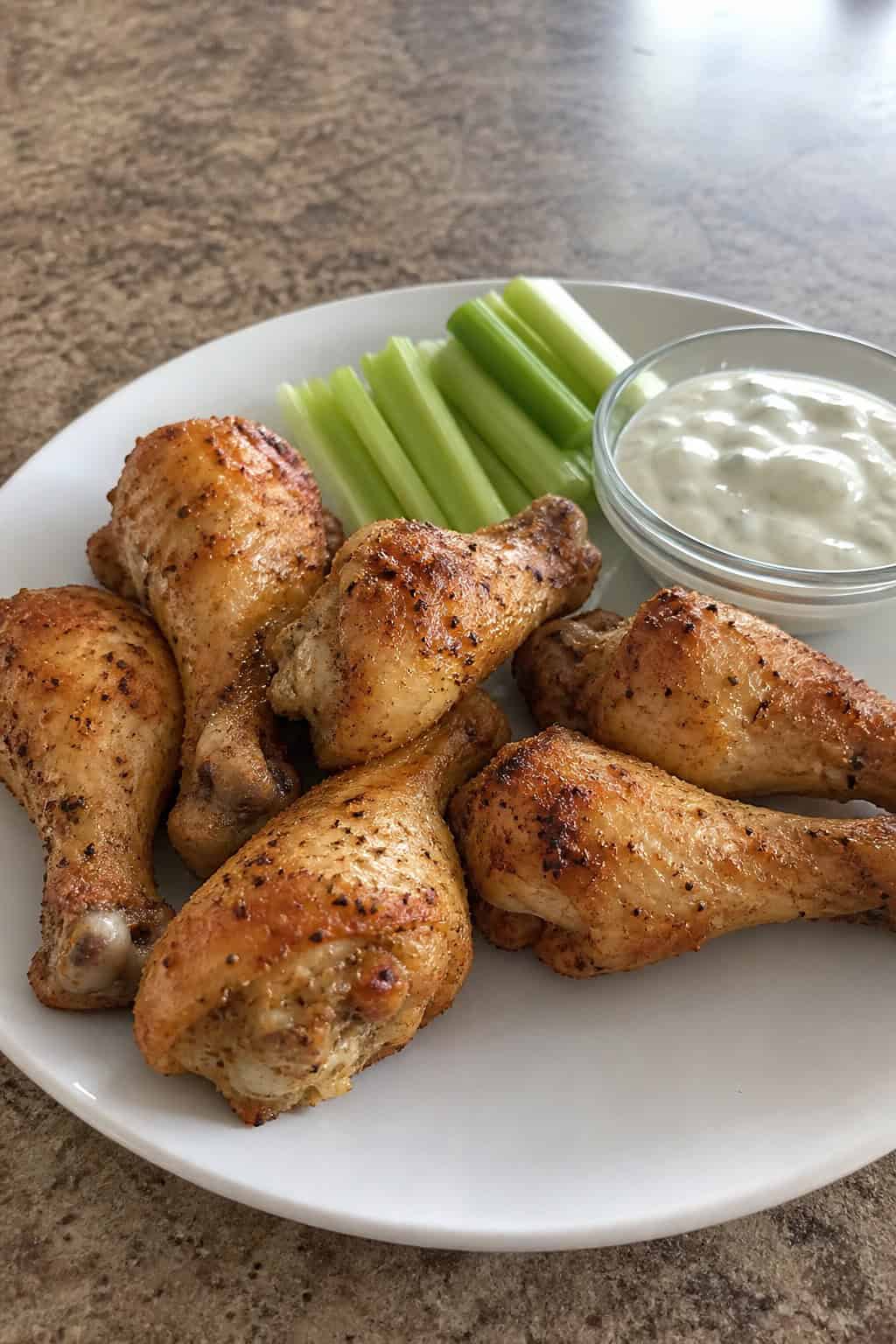 Crispy air-fried chicken drumsticks on a white plate next to celery sticks and a small bowl of creamy ranch dipping sauce on a light kitchen countertop.