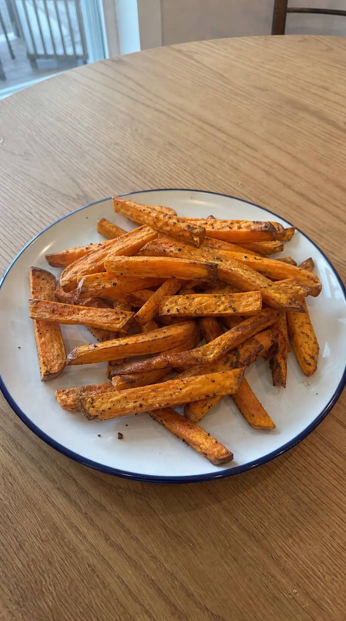 A bowl of golden air-fryer sweet potato fries on a wooden table with fresh sweet potatoes and herbs around.