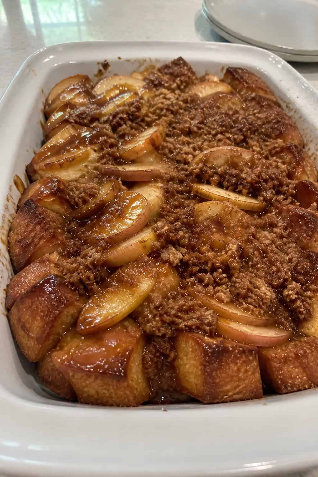 A baked French toast casserole topped with caramelized apple slices and a golden streusel crust in a cream ceramic dish on a wooden table.